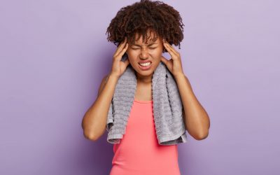 Exhausted tired dark skinned woman wears pink tanktop, touches temples, suffers from headache after cardio training, frowns face from bad pain, has towel around neck, isolated on purple background