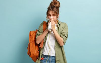 People, disease concept. Ill dark haired woman sneezes in handkerchief, carries rucksack, listens music in headphones, dressed casually, stands against blue background. Student feels unwell.